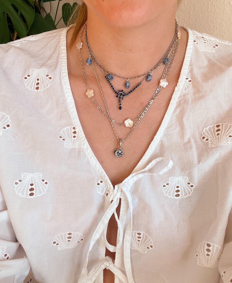May include: A collection of layered necklaces in silver tones. The necklaces feature various pendants, including floral and gemstone designs. The model is wearing a white blouse with a seashell pattern.
