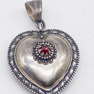 May include: A silver heart pendant with a decorative border and a small red gemstone. The pendant has a loop for a chain. The metal has a slightly tarnished appearance, suggesting an antique or vintage piece. The pendant is likely made of sterling silver.