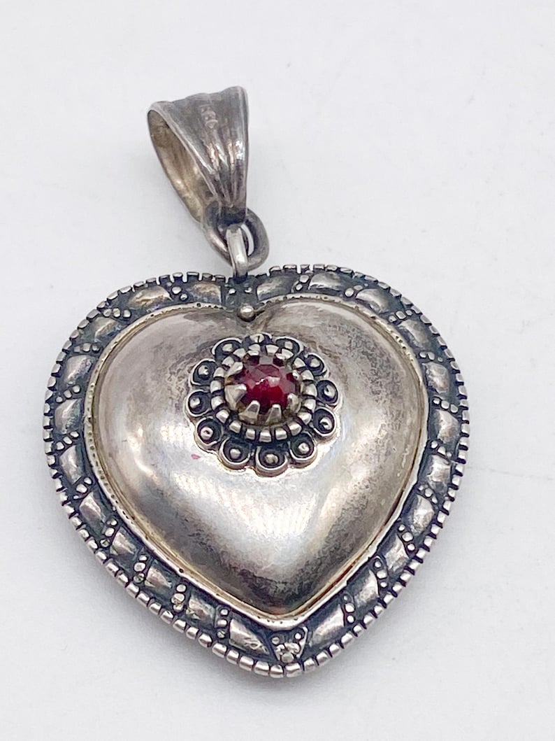 May include: A silver heart pendant with a decorative border and a small red gemstone. The pendant has a loop for a chain. The metal has a slightly tarnished appearance, suggesting an antique or vintage piece. The pendant is likely made of sterling silver.