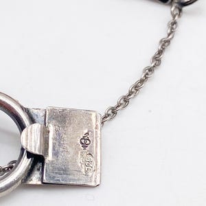 Puede incluir: Primer plano de un cierre y cadena de pulsera de plata. El cierre es rectangular con un cierre de bisagra y un peque&ntilde;o lazo. La cadena es delicada y el metal tiene un acabado pulido. El cierre est&aacute; estampado con marcas.