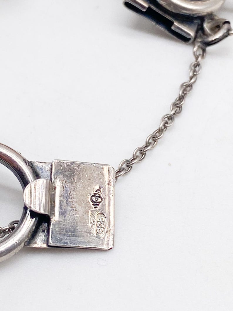 Puede incluir: Primer plano de un cierre y cadena de pulsera de plata. El cierre es rectangular con un cierre de bisagra y un peque&ntilde;o lazo. La cadena es delicada y el metal tiene un acabado pulido. El cierre est&aacute; estampado con marcas.