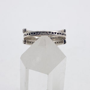 May include: A silver ring with a double band design. The top band features a row of small hearts, while the bottom band has a row of small dots. The ring is sitting on a clear crystal.