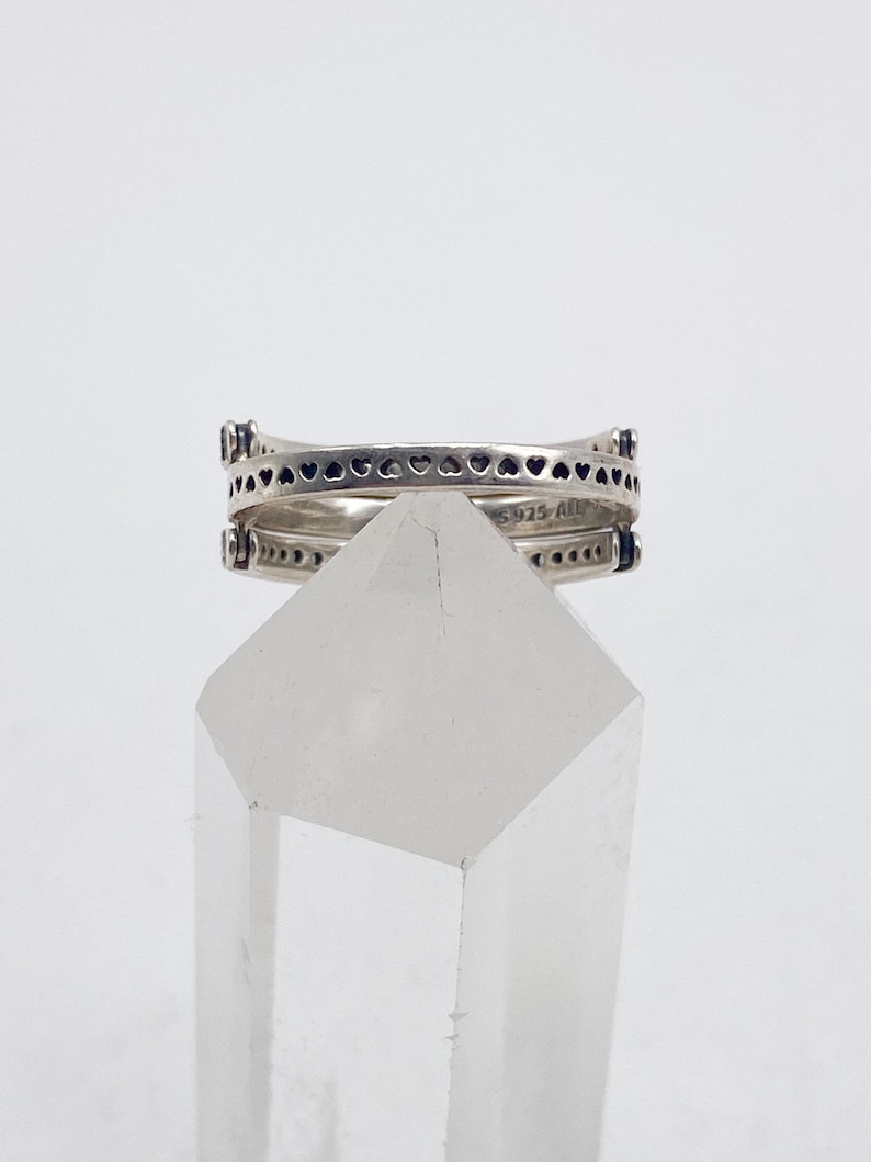 May include: A silver ring with a double band design. The top band features a row of small hearts, while the bottom band has a row of small dots. The ring is sitting on a clear crystal.