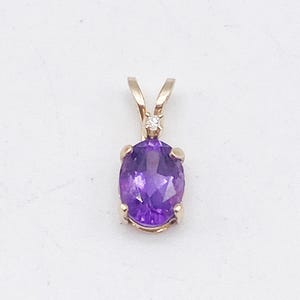 May include: A gold pendant featuring an oval-cut, deep purple amethyst gemstone. A small diamond is set above the amethyst. The pendant has a simple, elegant design, suitable for everyday wear or special occasions. The gemstone is approximately 0.5 inches in length.