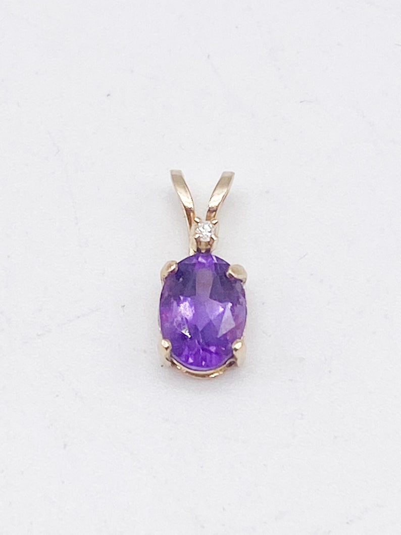 May include: A gold pendant featuring an oval-cut, deep purple amethyst gemstone. A small diamond is set above the amethyst. The pendant has a simple, elegant design, suitable for everyday wear or special occasions. The gemstone is approximately 0.5 inches in length.