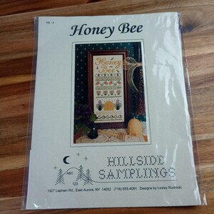 May include: A cross-stitch pattern titled "Honey Bee" by Hillside Samplings. The design features a framed image with the words "Honey Bee" and a beehive. The pattern is packaged in a clear plastic sleeve.