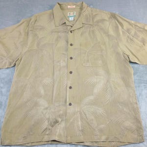 May include: A light tan button-down shirt with a palm tree pattern. The short-sleeved shirt has a chest pocket and a classic collar. The palm tree design is a lighter shade of tan, creating a subtle contrast. The shirt is made of a lightweight fabric.