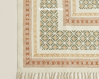 May include: A cream-colored rug with a geometric pattern in shades of green, brown, and beige. The rug has a fringe border.