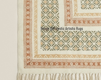 May include: Cream-colored rug with geometric patterns in shades of brown, tan, and green. The rug features a diamond pattern, bordered by a series of circles and triangles. The bottom edge has a fringe.