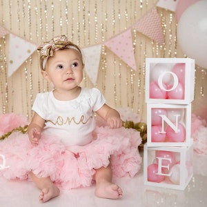 May include: A baby girl wearing a pink tutu and a white shirt with gold glitter lettering that says "one". She is wearing a gold and pink bow headband.  Behind her are white boxes with pink balloons inside. The boxes spell out "ONE".
