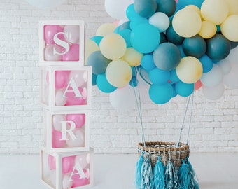 May include: Four white boxes stacked on top of each other with the letters S, A, R, and A inside. The boxes are filled with pink and white balloons. A hot air balloon made of balloons in various colors, including blue, pink, white, and yellow, is in the background.