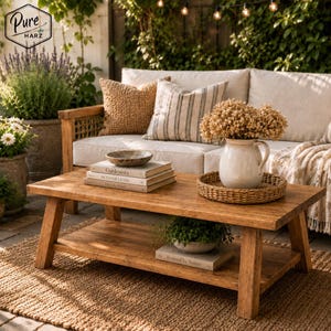 Könnte beinhalten: Ein rustikaler Couchtisch aus Holz mit einem unteren Regal, auf dem eine Topfpflanze und ein Buch stehen. Der Tisch steht auf einem gewebten Teppich, im Hintergrund ein Sofa und Kissen. Ein weißer Krug mit getrockneten Blumen steht auf einem gewebten Tablett.