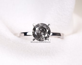 Puede incluir: Un anillo de compromiso solitario con un gran diamante redondo de sal y pimienta engastado en una banda de plata pulida. El diamante tiene un aspecto moteado y gris. El anillo se muestra sobre un fondo de tela blanca.