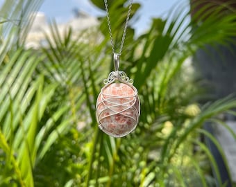 May include: A silver pendant necklace featuring a large, oval sunstone wrapped in delicate silver wire. The sunstone has a speckled, peach and orange color. The necklace hangs against a backdrop of green foliage.
