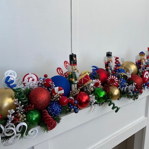 Puede incluir: Una guirnalda navideña con adornos rojos, verdes, dorados, azules y blancos, bastones de caramelo y figuras de cascanueces. La guirlande está decorada con cintas rizadas blancas y verdes.