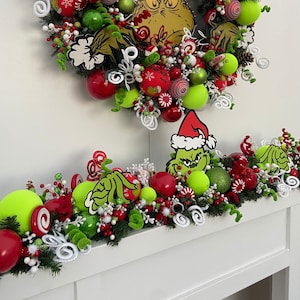 Puede incluir: Una corona y guirnalda navideña con el tema del Grinch. La corona está adornada con adornos rojos y verdes, esferas verde lima y detalles blancos. La guirnalda refleja el diseño de la corona, creando una exhibición festiva.