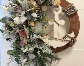 May include: A white anchor with a rope wrapped around it is attached to a brown wooden wreath. The wreath is decorated with green leaves, white and brown seashells, and starfish.
