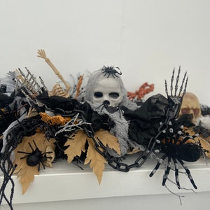 May include: A Halloween centerpiece featuring a white skull with black eyes, surrounded by black spiders, a skeleton, black and brown fabric, and a black chain.