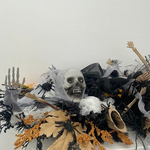 May include: Halloween centerpiece featuring a white skull, black spider webs, black and gold leaves, and black spiders. The centerpiece is on a white surface.