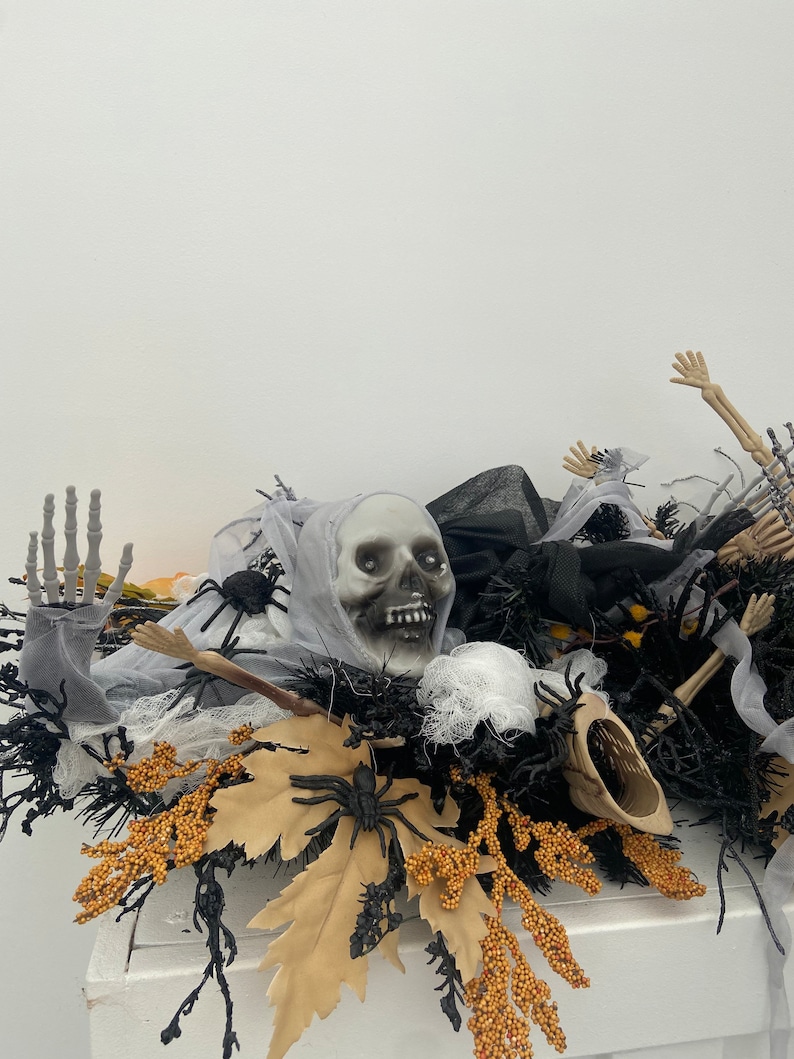 May include: Halloween centerpiece featuring a white skull, black spider webs, black and gold leaves, and black spiders. The centerpiece is on a white surface.