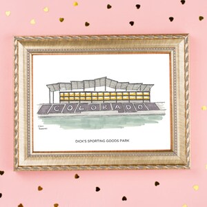 K&ouml;nnte beinhalten: Eine gerahmte Illustration eines Stadions mit dem Wort "COLORADO" in gro&szlig;en Buchstaben auf der Vorderseite. Der Text unter dem Stadion lautet "DICK'S SPORTING GOODS PARK".