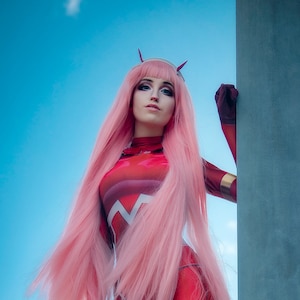 Puede incluir: Una mujer con un traje rojo y blanco, cabello rosa y cuernos posa contra un pilar gris. El fondo es un cielo azul con nubes blancas.