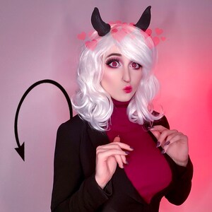 May include: A person in a cosplay outfit with white hair, black horns, and a black blazer over a burgundy turtleneck. Pink heart-shaped filters are around the head. A black devil tail is drawn on the wall.