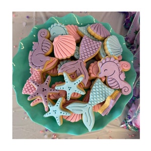May include: A turquoise serving dish filled with mermaid-themed biscuits. The biscuits are decorated in pastel colours, including pink, purple, and blue, and shaped like seashells, starfish, and seahorses. The biscuits are arranged on a turquoise plate.