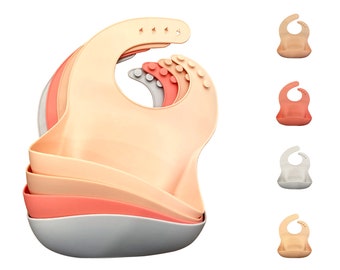 Puede incluir: Un juego de cuatro baberos de silicona en diferentes colores: melocotón, coral, gris y azul claro. Los baberos están apilados uno encima del otro, con el babero de melocotón en la parte superior. Los baberos tienen una forma redondeada y una abertura para el cuello.