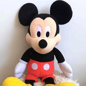 May include: A plush Mickey Mouse doll, featuring black ears, a black body, red shorts with white circles, and large yellow shoes. The doll has a tan face, black eyes, and white gloves. It is sitting on a fluffy, white surface.