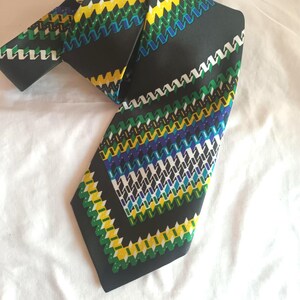 Pode incluir: Uma gravata preta com um padrão geométrico repetitivo em amarelo, verde, azul e branco. A gravata é feita de um tecido liso e é exibida em uma superfície branca. O design é uma série de ziguezagues.
