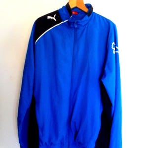 Puede incluir: Una chaqueta deportiva azul brillante con cremallera y paneles negros en los hombros y los costados. La prenda incluye un cuello alto, una cremallera frontal completa y logotipos de Puma en blanco en el hombro derecho y la manga izquierda.