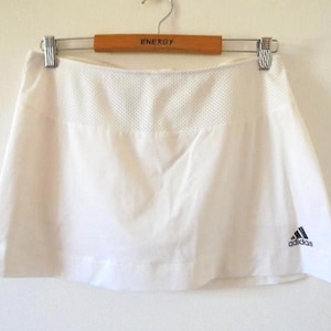 May include: White athletic skirt with a mesh panel at the waist, hanging on a wooden hanger. The Adidas logo is visible on the lower right. The skirt is designed for sports activities.