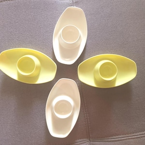 May include: Four egg cups, two in a pale yellow and two in white. Each cup has an oval base with a circular indentation to hold an egg. The cups are arranged on a gray surface.