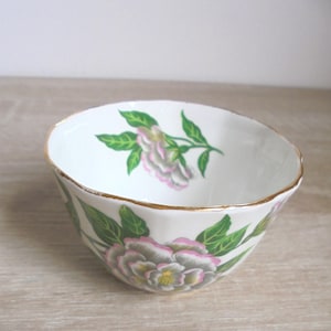 May include: White porcelain bowl with a floral design. The bowl features a gold rim and is decorated with pink, gray, and green flowers and leaves. The bowl is likely for serving or display.
