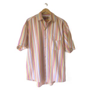 Puede incluir: Una camisa de manga corta con botones y un patrón de rayas verticales en tonos amarillo, rosa, blanco y morado claro. La camisa incluye un bolsillo en el pecho en el lado izquierdo y un cuello, con una etiqueta que dice SC51 JEANS.