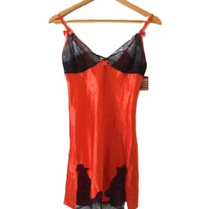 Può includere: Un vestito sottoveste in raso rosso con dettagli in pizzo nero. L'abito presenta un corpetto e un orlo in pizzo nero, con piccoli fiocchi sulle spalline. L'indumento è appeso a una gruccia di legno su sfondo bianco.