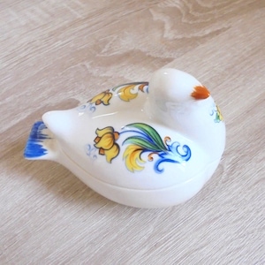 May include: A white ceramic bird-shaped trinket box with a lid. The bird features yellow, blue, and green floral patterns, a blue tail, and an orange beak. It rests on a light brown wooden surface.