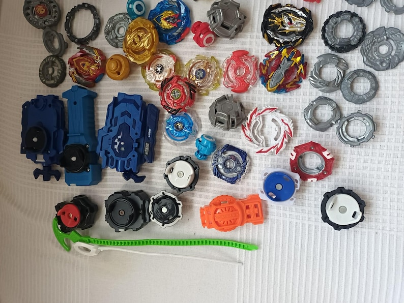 Op de afbeelding: Een verzameling kleurrijke tol speelgoed en lanceerders. De tops hebben verschillende ontwerpen en kleuren, waaronder rood, blauw, goud en zilver. De lanceerders zijn voornamelijk blauw en zwart, met groene trekkoorden. Het speelgoed is gerangschikt op een wit oppervlak.