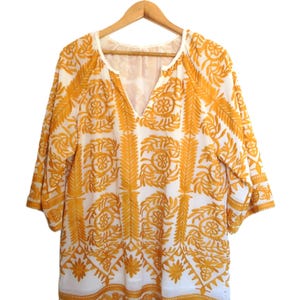 Op de afbeelding: Witte tuniek met een V-hals en driekwart mouwen, versierd met ingewikkelde gele borduursels. De borduursels tonen bloemen- en geometrische patronen, wat een bohemien uitstraling creëert. De tuniek hangt aan een houten hanger.