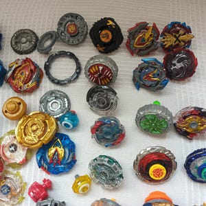 Op de afbeelding: Een verzameling kleurrijke tolletjes in verschillende ontwerpen en maten. De tolletjes hebben een mix van rode, blauwe, gouden en zilveren accenten. Sommige tolletjes hebben ingewikkelde patronen en gelaagde componenten, met launchers links.
