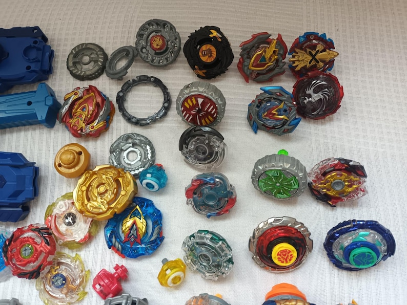 Op de afbeelding: Een verzameling kleurrijke tolletjes in verschillende ontwerpen en maten. De tolletjes hebben een mix van rode, blauwe, gouden en zilveren accenten. Sommige tolletjes hebben ingewikkelde patronen en gelaagde componenten, met launchers links.