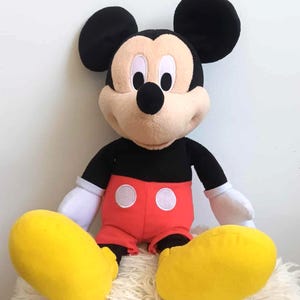 May include: A plush Mickey Mouse doll with black ears, a black body, red shorts with white buttons, and large yellow feet. The doll has a tan face, white gloves, and is sitting on a white, fluffy surface.