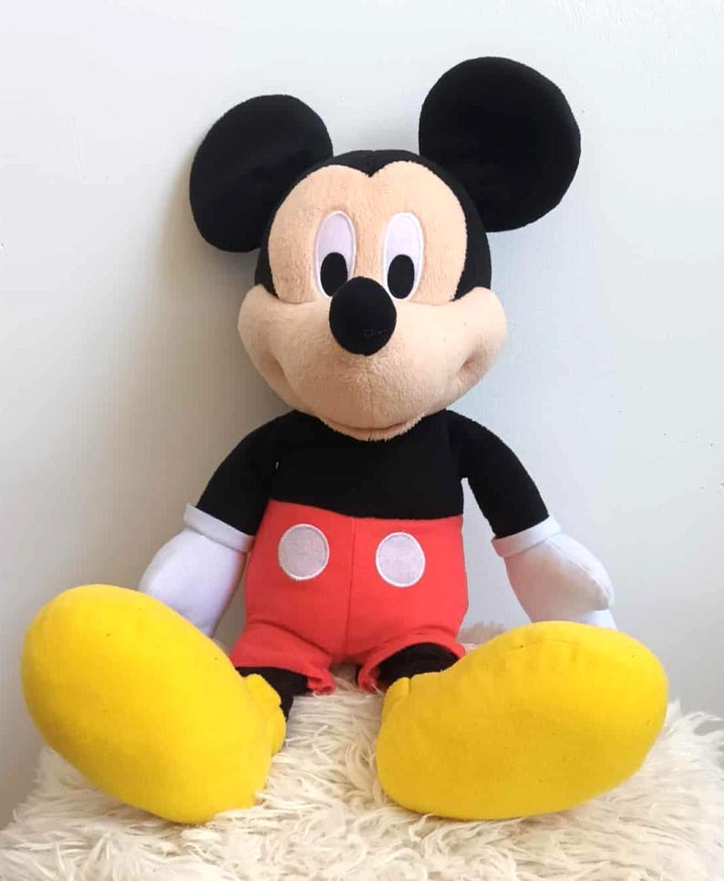 May include: A plush Mickey Mouse doll with black ears, a black body, red shorts with white buttons, and large yellow feet. The doll has a tan face, white gloves, and is sitting on a white, fluffy surface.