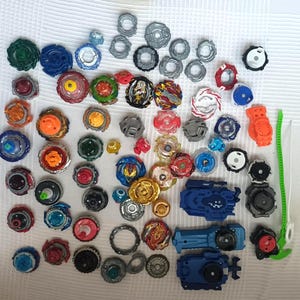Op de afbeelding: Een verzameling kleurrijke tollen en lanceerders in verschillende maten en ontwerpen. De tollen hebben een verscheidenheid aan kleuren, waaronder blauw, rood, groen en goud. Er zijn ook verschillende lanceerders zichtbaar.