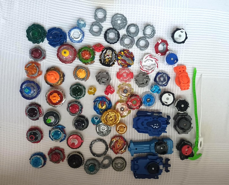 Op de afbeelding: Een verzameling kleurrijke tollen en lanceerders in verschillende maten en ontwerpen. De tollen hebben een verscheidenheid aan kleuren, waaronder blauw, rood, groen en goud. Er zijn ook verschillende lanceerders zichtbaar.
