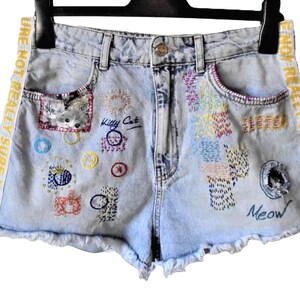 Könnte beinhalten: Hellblaue Jeansshorts mit ausgefransten Säumen, hoher Taille und Seitenstreifen mit dem Text NOT REALLY SURE NOT REALLY. Die Vorderseite zeigt bunte Stickmuster, eine kleine Katzengrafik sowie die Wörter Kitty Cat und Meow.