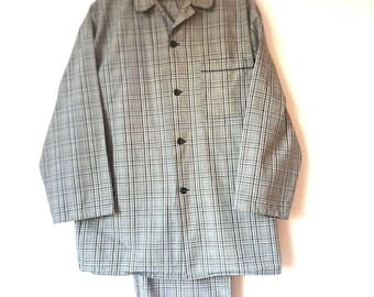 Puede incluir: Un conjunto de pijama a cuadros grises y blancos, que incluye una camisa con botones y bolsillo en el pecho y pantalones a juego. El pijama está colgado de una percha de madera sobre un fondo blanco. La camisa tiene botones y ribetes negros.
