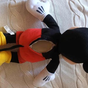 May include: A Mickey Mouse plush toy, lying on its back, features a black head and body, red shorts, yellow shoes, and white gloves. The toy has a visible opening in the back. The plush is on a cream-colored knitted surface.