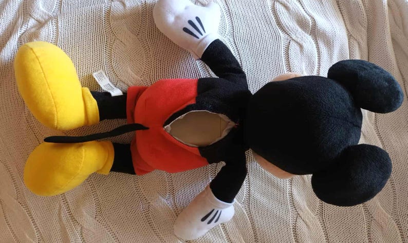 May include: A Mickey Mouse plush toy, lying on its back, features a black head and body, red shorts, yellow shoes, and white gloves. The toy has a visible opening in the back. The plush is on a cream-colored knitted surface.